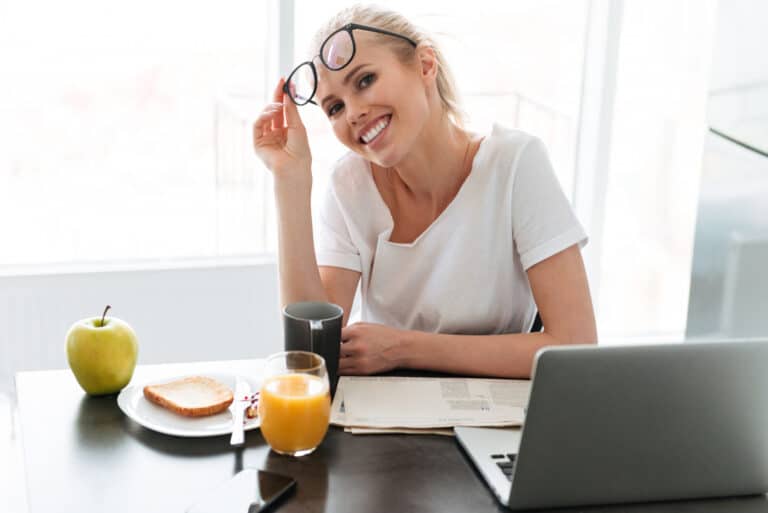 Uśmiechnięta kobieta przy biurku z laptopem i kawą, zdejmująca okulary podczas pracy przy komputerze – przykład codziennych nawyków pokazujących, jak dbać o wzrok w domowych warunkach.